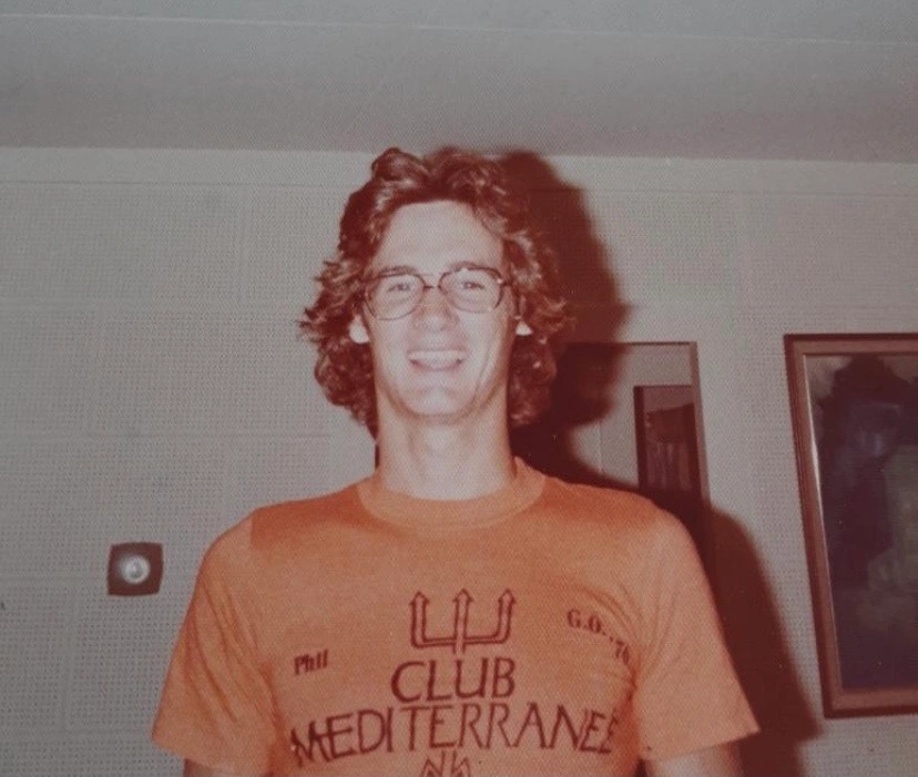 Young Phil Bennett smiling with his partner in front of lush green foliage, wearing a casual rock and roll t-shirt