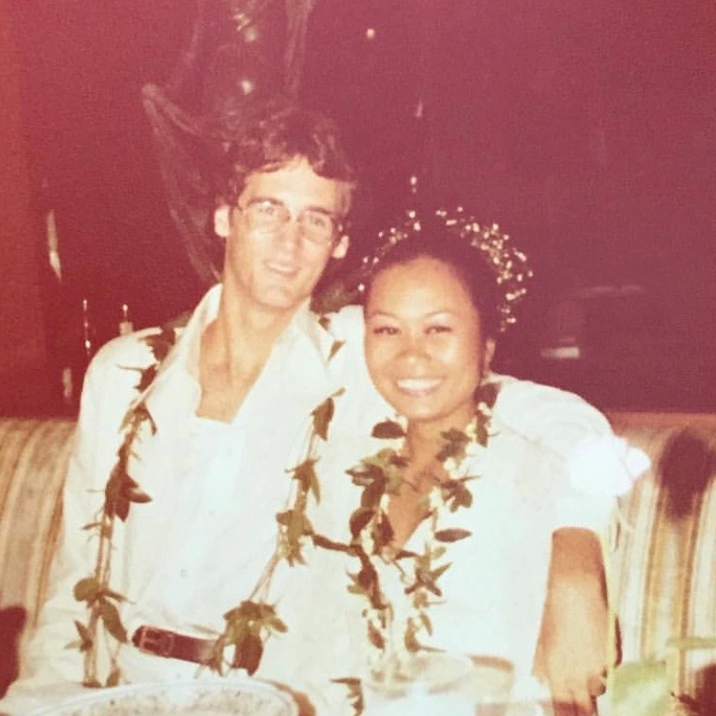 A young Phil Bennett in a white suit standing with his partner in a red dress, smiling by a scenic coastal or lake backdrop
